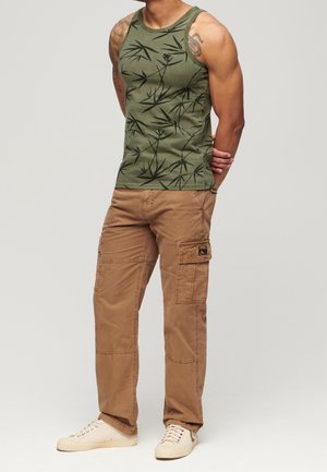 Grünes Tanktop mit schwarzem Blättermuster, kombiniert mit beigefarbenen Cargo-Hosen mit Taschen und beigen Sneakers mit strukturierter Oberfläche.