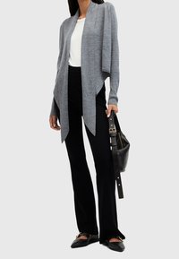 Szary kardigan z odkrytym przodem i długimi rękawami, noszony na białym topie, zestawiony z czarnymi spodniami o rozszerzanych nogawkach i czarnymi płaskimi butami. Torba na ramię.
