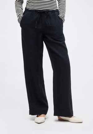 Άτομο φοράει μαύρο παντελόνι με φαρδιά μπατζάκια, ελαστική μέση και κορδόνι, σε συνδυασμό με ριγέ μακρυμάνικη μπλούζα και άσπρα φλατ παπούτσια.