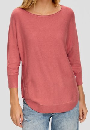 Femme portant un pull ample à manches longues couleur vieux rose avec un large col et un jean bleu clair déchiré.