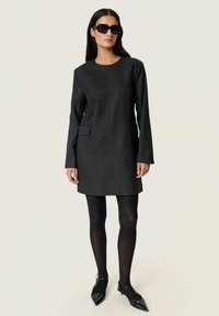 Schwarzes Nadelstreifenkleid mit langen Ärmeln, runder Ausschnitt und Seiten Taschen. Kombiniert mit schwarzen Strumpfhosen und spitzen Ballerinas. Dunkle Sonnenbrille.