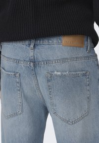 Světle modré džíny z denimu s vybledlým povrchem, s dvěma zadními kapsami a hnědou koženou nášivkou značky na pase.