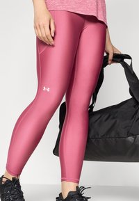 Różowe legginsy sportowe o gładkiej, błyszczącej teksturze z kontrastowym białym logo na udzie; w połączeniu z czarną torbą na siłownię.