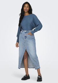 Jupe maxi en denim avec une fente avant, taille haute et un délavage bleu classique. Associée à un pull bleu texturé et des chaussures noires épaises.