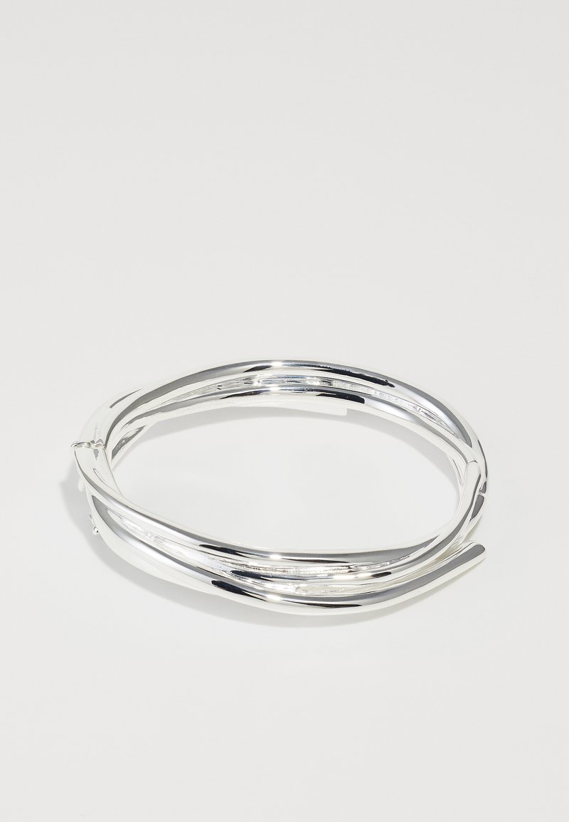 Sudraba metāla aproce ar savijušu dizainu, gludu tekstūru un spīdīgu apdari. Raksturīga plāna, elastīga struktūra, kas nodrošina ciešu piegulšanos.