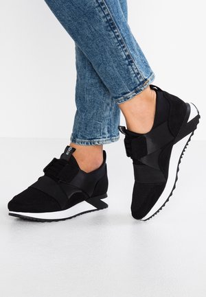 Henkilö, joka pukeutunut sinisiin farkkuihin ja mustiin slip-on-tennareihin, joissa leveät kuminauhat ja valkoiset pohjat, seisoo valkoisella lattialla.