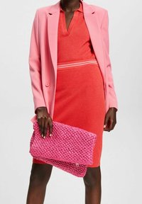 Vaaleanpunainen bleiseri punaisen, raidallisen yksityiskohdan omaavan mekko päällä, pitäen kädessään teksturoitua vaaleanpunaista clutch-laukkua. Bleiseri on muotoiltu hyvin; mekko on istuva.