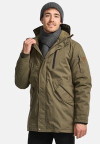 Khaki parka jakke med lynlås foran, justerbar hætte, sidelommer og ribbede manchetter. Bæres med et gråt tørklæde og en hue.