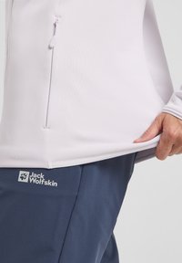 Camicetta lunga manica di colore viola chiaro con zip laterale e texture liscia, abbinata a pantaloni blu navy con un logo sulla coscia.