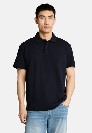 Man met kort zwart haar, gekleed in een zwart polohemd met korte mouwen en lichtblauwe spijkerbroek tegen een effen achtergrond.