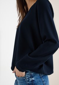 Blouse bleu marine à manches longues en tissu lisse, avec un décolleté en V et une coupe décontractée, associée à un jean bleu clair déchiré.