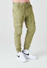 Pantalones cargo verde claro con cintura elástica, cordón, bolsillos laterales y puños acanalados; combinados con zapatillas blancas con detalles en negro.