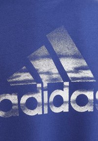 Sininen t-paita, jossa on suuri valkoinen teksturoitu adidas-logo, sileä puuvillapinta ja pyöreä kaula-aukko.