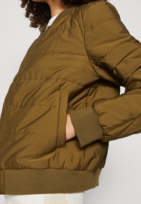 Personne portant une veste bomber matelassée vert olive avec poignets et ourlet côtelés, main dans la poche latérale, sur un fond uni.
