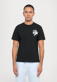 Schwarzes T-Shirt mit kurzen Ärmeln aus Baumwolle, mit einem bunten Grafikdesign auf der linken Brust. Das Model trägt hellblaue gestreifte Hosen.