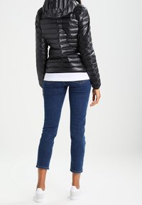Chaqueta acolchada negra con acolchado horizontal, diseño ajustado, capucha incorporada. Combinada con jeans azules y zapatillas blancas, mostrando un atuendo casual.