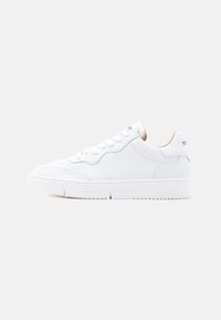 Λευκό sneaker από δέρμα με χαμηλή γραμμή, κορδόνια στο μπροστινό μέρος και εσωτερική επένδυση σε μπεζ, εμφανίζεται σε πλευρική όψη σε απλό φωτεινό φόντο.