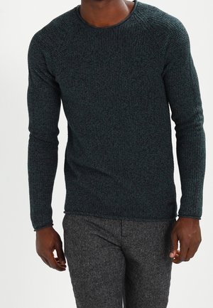 Man draagt een donkergroene, getextureerde, lange mouwen trui en grijze wollen broek, staand voor een effen achtergrond.