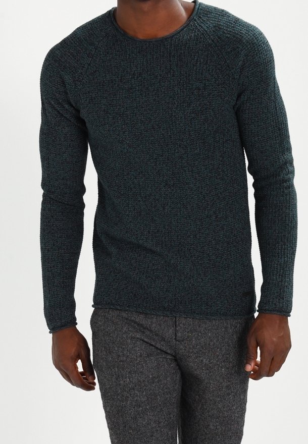 Homme portant un pull à manches longues texturé vert foncé et un pantalon en laine gris, debout devant un fond uni.