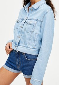 Persona vistiendo una chaqueta de mezclilla azul claro con detalles de botones y pantalones cortos de mezclilla azul oscuro arremangados frente a un fondo blanco sencillo.