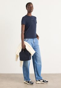 T-shirt di cotone navy a maniche corte, abbinato a jeans dritti blu chiaro. Tiene in mano una borsa scura con texture e un maglione crema.