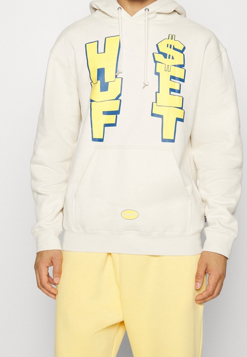 Personne portant un sweat à capuche écru avec des lettres "HUF" jaune vif et bleu, ainsi qu'un signe dollar, combiné à un pantalon jaune clair.