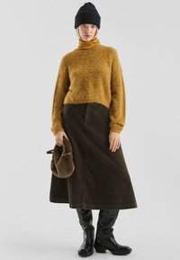 Mosterdkleurige fuzzy trui, bruine corduroy midi rok, zwarte kniehoge laarzen en een bruine bont clutch. Een zwarte beanie maakt de outfit compleet.