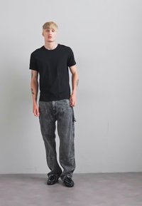 Un jeune homme aux cheveux blonds se tient contre un mur gris, portant un t-shirt noir, un pantalon cargo gris ample et des chaussures noires, avec des tatouages visibles sur les bras.
