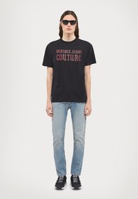 T-shirt nera in cotone con la scritta "Versace Jeans Couture" in lettere rosse, abbinata a jeans blu chiaro e sneakers nere. Occhiali da sole moderni indossati.