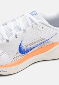Chaussure de sport blanche avec le logo Nike bleu, semelle intermédiaire orange, lacets blancs et dessus en mesh, montrant le côté et une partie de la semelle.
