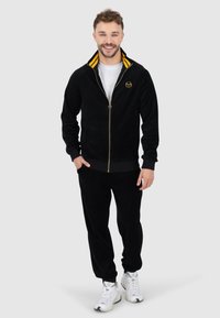Schwarzer Velour-Sportanzug mit Reißverschlussjacke, gelb gestreifter Kragen, goldfarbenem Logo auf der Brust und passenden Hosen. Weiße Sneakers runden das Outfit ab.