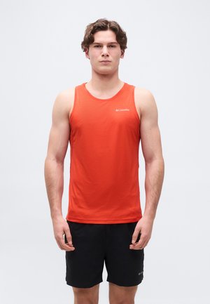 Mladić stoji okrenut prema naprijed, nosi narančastu majicu bez rukava i crne kratke hlače, na jednobojnoj bijeloj pozadini.