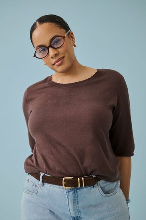 Frau mit Brille, die einen braunen Pullover und hellblaue Jeans trägt, steht vor einem einfarbigen hellblauen Hintergrund.
