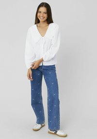 Blouse blanche avec un col festonné et un lien à l'avant, associée à un pantalon en denim bleu décoré de petits motifs animaliers. Des chaussures décontractées complètent le look.