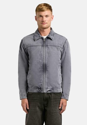 Mann trägt eine verblasste graue Reißverschlussjacke über einem weißen Hemd und dunkler Hose, steht vor einem einfarbigen hellen Hintergrund.