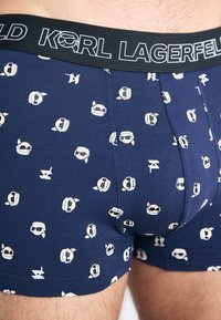 Tengerész kék boxer alsónemű, fekete elasztikus derékpánttal, amelyen fehér "KARL LAGERFELD" felirat található. Kicsi, játékos grafikus fejmotívumokkal van mintázva.
