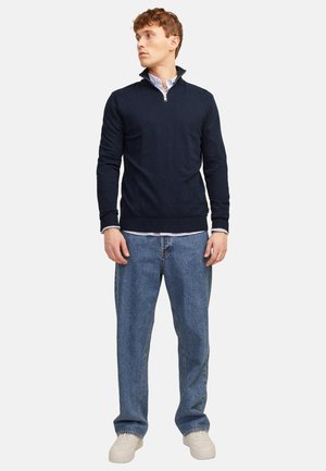 Mann trägt einen marineblauen Pullover mit einem Viertel-Reißverschluss über einem weißen Hemd, lockere blaue Jeans und weiße Sneaker, steht vor einem schlichten hellen Hintergrund.