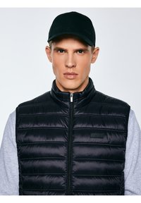 Fiatal férfi fekete baseballsapkában és fekete steppelt mellényben, világos szürke hosszú ujjú ing fölött, előre néz semleges arckifejezéssel.