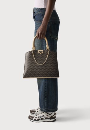 Sac à main marron à motif avec des poignées beiges et un accent en chaîne, tenu par une personne portant un jean bleu et des baskets marron et blanches.