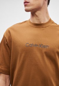 Καφέ βαμβακερό T-shirt με στρογγυλή λαιμόκοψη και κοντά μανίκια. Διαθέτει λογότυπο "Calvin Klein" κεντημένο σε γκρι στο στήθος.