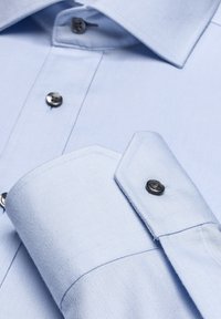 Hellblaues Button-up-Hemd mit strukturiertem Kragen, schwarzen Knöpfen und glatter Textiloberfläche. Die Manschette hat ein scharfes, abgeschrägtes Design.