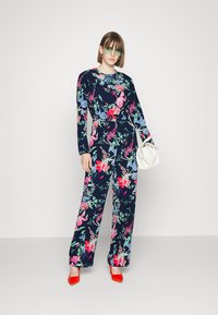 Macacão florido azul-marinho com mangas compridas, apresentando flores rosas e azuis. Combinado com sapatos de salto vermelhos e uma mala branca.