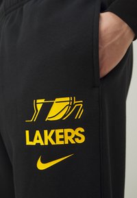 Sorte joggebukser med gule "LAKERS"-bokstaver og logo, laget av glatt stoff, med sidelommer og en Nike-svung ved kanten.