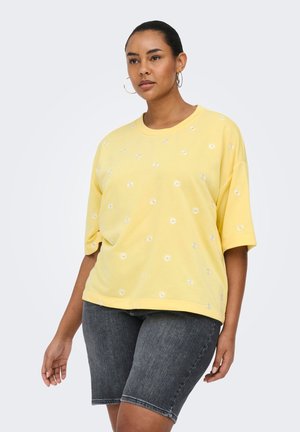 Frau trägt ein gelbes oversized T-Shirt mit kleinen weißen Gänseblümchen und dunkelgrauen Denim-Bermuda-Shorts, steht vor einem einfarbigen Hintergrund.