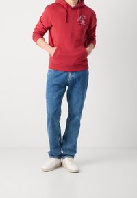Felpa rossa con cappuccio e tasca frontale, abbinata a jeans di denim blu e sneakers bianche. La felpa presenta un design grafico sul petto.