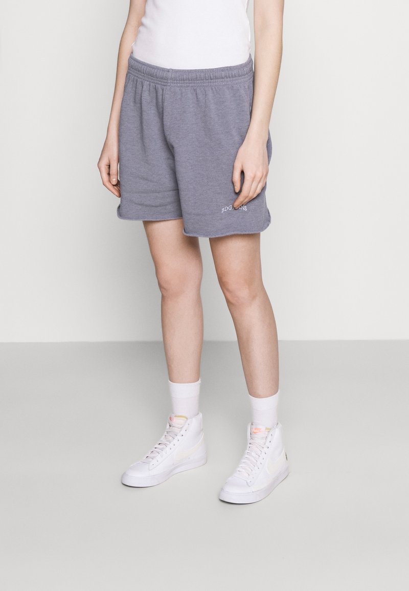 Šedé bavlněné kraťasy s elastickým pasem, doplněné bílým logem. V kombinaci s bílými vysokými teniskami a bílými ponožkami, umístěné na jednobarevném pozadí.