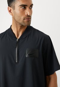 Sort Nike polo-shirt med lynlås krave, med struktureret stof og et sort gummilogo på brystet.
