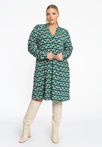 Robe verte à motifs avec des manches longues et un décolleté en V, associée à des bottes beige montantes. Le design comprend des formes géométriques.