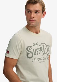 Camiseta beige claro de algodón con cuello redondo. Presenta un logo impreso en gris oscuro y una pequeña etiqueta roja en la manga.