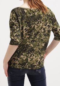 Oliivinvihreä maastopaita, jossa on 3/4-hihat, pyöreä kaula-aukko ja pehmeä kangas. Yhdistetty tummansinihin farkkuihin, joilla on rento istuvuus.